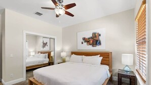 A hotel room with a large bed, a ceiling fan, a wall-mounted artwork, a bedside table with a lamp, and a window with blinds. - Mesa House Dos - One block from Duval St (Key West)