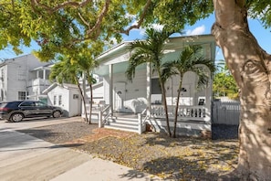 A white house with a porch, palm trees, and a car parked in front.