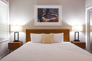 Une chambre d’hôtel avec un grand lit, deux tables de chevet avec des lampes, un tableau encadré au mur et une fenêtre avec des stores.