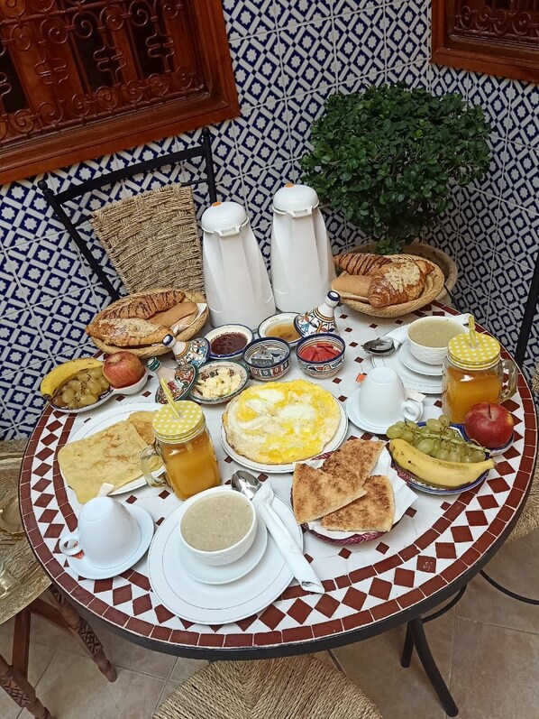 A round table with a patterned tablecloth, various breakfast items including bread, fruits, and beverages, and a decorative wall with a geometric pattern.