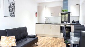 Una cocina moderna con armarios blancos, encimera negra y electrodomésticos de acero inoxidable. En la zona de estar, hay un sofá de cuero negro con un cojín estampado.