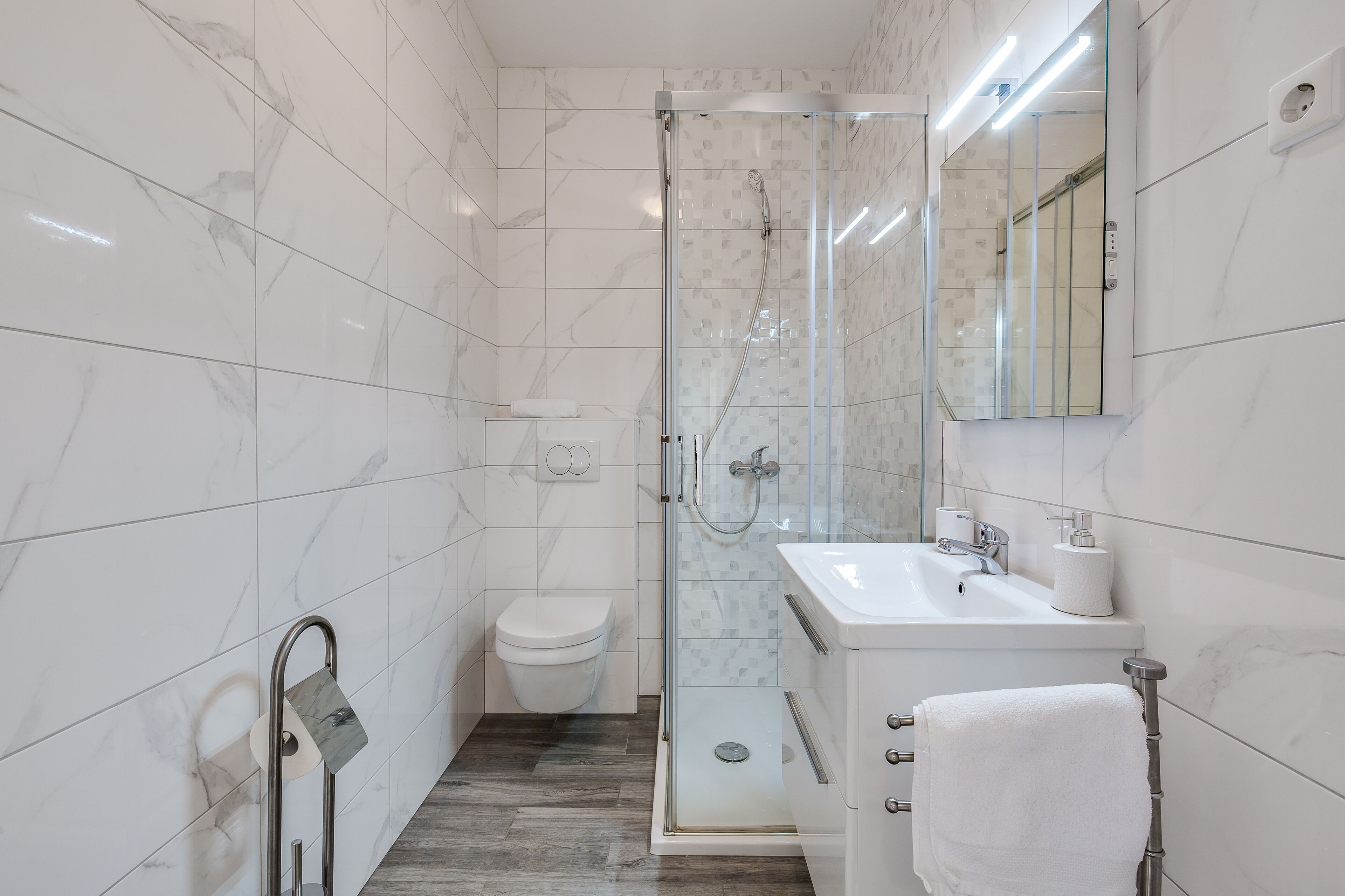Un bany modern amb plat de dutxa envoltat de vidre, un vàter blanc i un lavabo amb barra de tovalloles.
