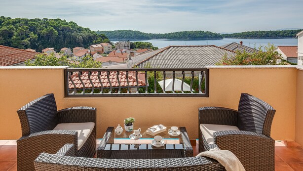 A balcony with wicker furniture, a glass table, and a view of a lake and town. - JURIĆ apartments with outdoor pool and bargain in RAB-PALIT (Palit)