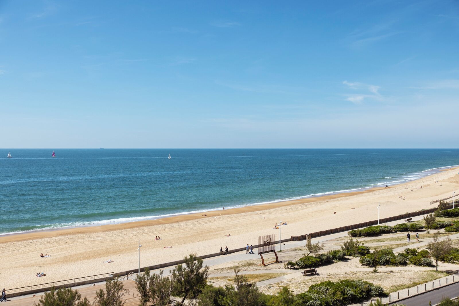Ein Strand mit sandigem Ufer, einer Promenade und blauem, wolkenlosem Himmel.