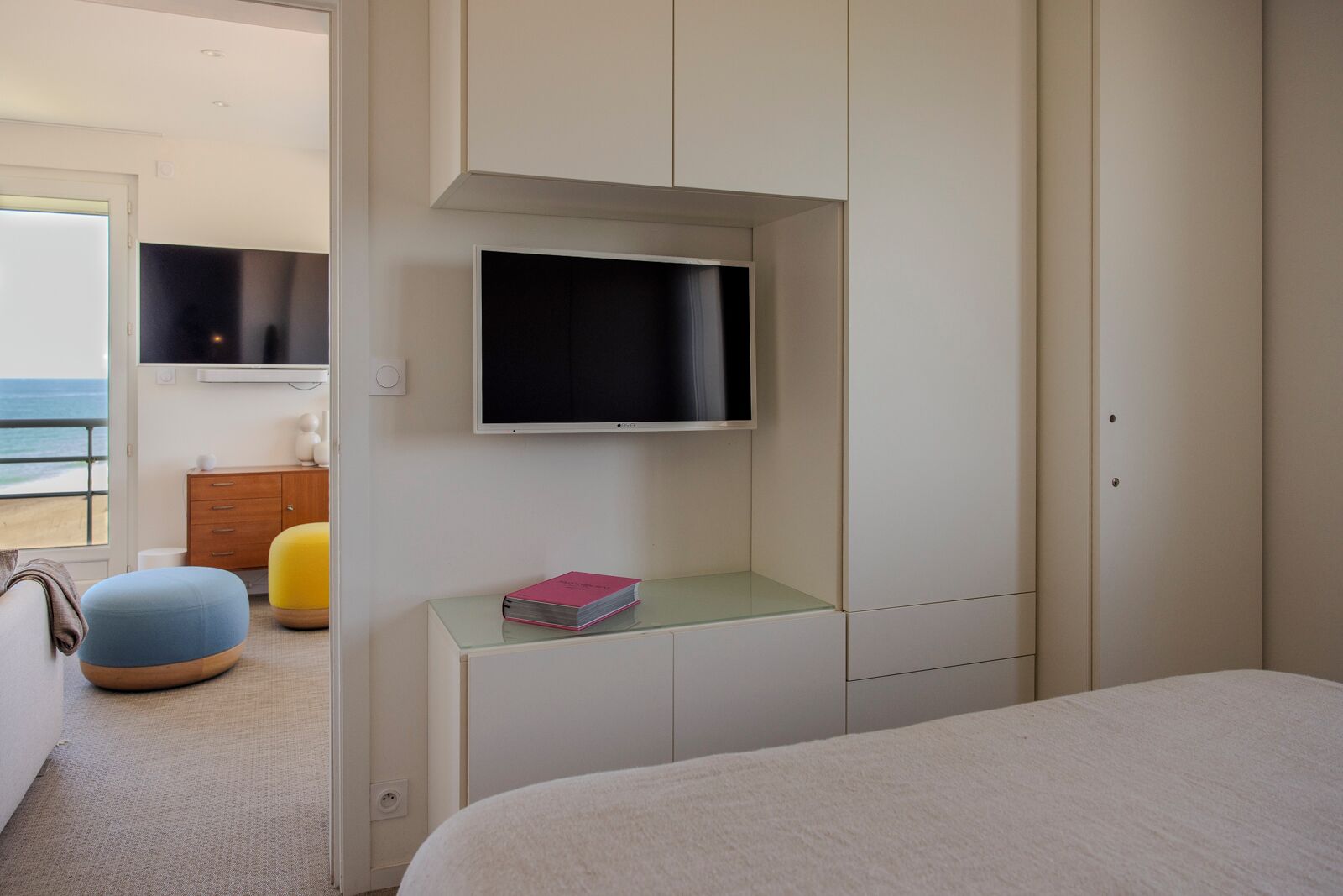 A bedroom with a bed, a TV mounted on the wall, a small bedside table with a book, and a view of the beach through an open door.