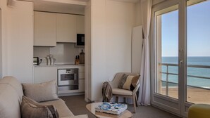 A modern living room with a sofa, armchair, and a small table with a book. A kitchen is visible in the background with an oven and microwave. The room has a large sliding glass door leading to a balcony with a view of the ocean. - Superb view of the ocean, the beach at your feet! (Soorts-Hossegor)