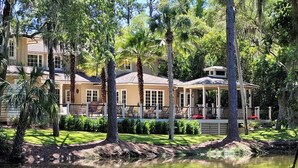 A large, multi-story house with a covered patio area, surrounded by lush greenery and a pond. - 48 S. Sea Pines Dr - Luxury Home in Sea Pines w/ Game Rm, Media Rm, Private Pool (Hilton Head Island)