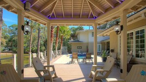 A covered outdoor deck with chairs, a pool, and a view of a house. - 48 S. Sea Pines Dr - Luxury Home in Sea Pines w/ Game Rm, Media Rm, Private Pool (Hilton Head Island)