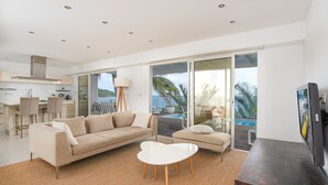 A modern living room with a sofa, a coffee table, and a TV. There is a kitchen area with a bar and a dining table. The room has large windows with a view of the ocean and palm trees. - Villa Leynita Bay, 5 bedrooms, sleeps 12 on the beach of Diamant Martinique  (le diamant)