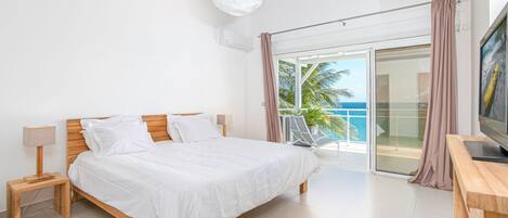 Une chambre avec un lit en bois, une télévision, une lampe et un balcon donnant sur l’océan.