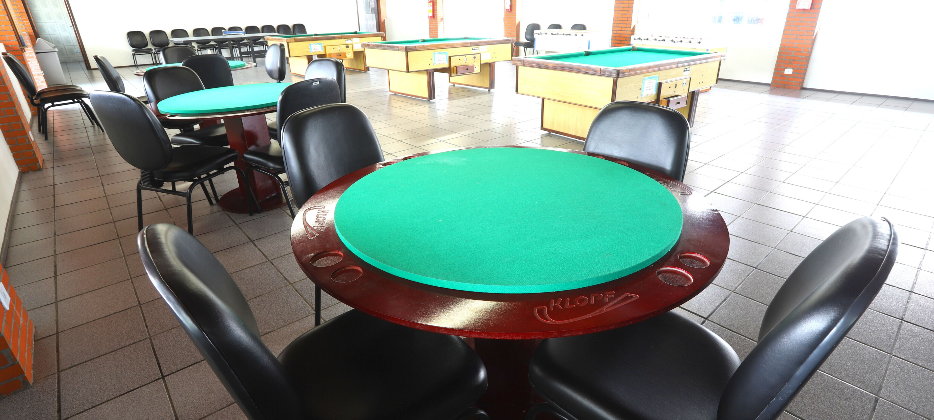 Una mesa de billar con superficie de fieltro verde, rodeada de sillas negras, en una sala con suelo de baldosas y varias mesas de billar.