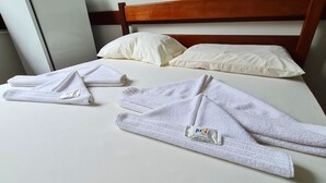 Un lit bien fait, avec du linge de lit blanc, comprenant deux serviettes et une paire de chaussons, dans une chambre dotée d’une tête de lit en bois et d’oreillers blancs.
