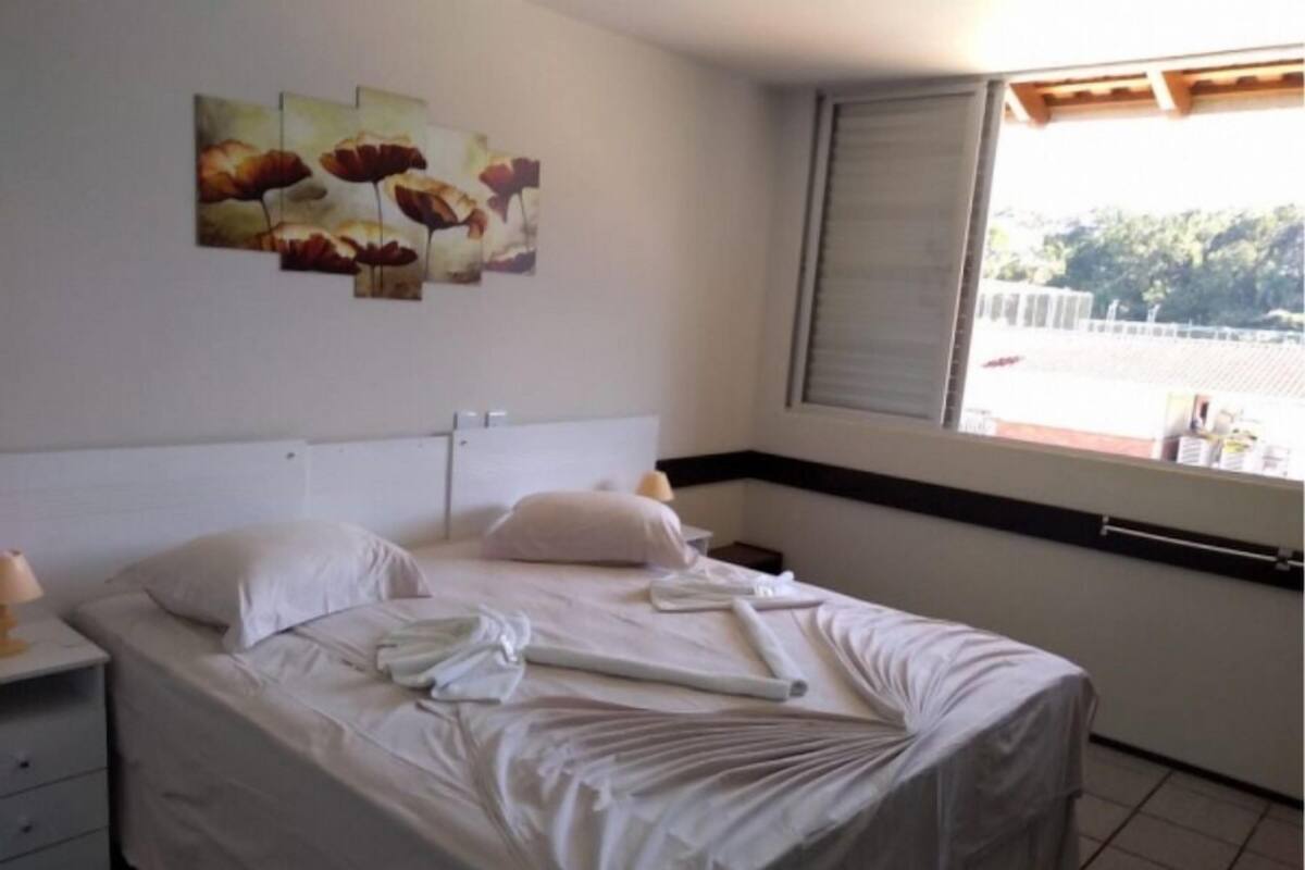 Cama bem arrumada com lençóis brancos, mesa de cabeceira com abajur e janela com vista para árvores.