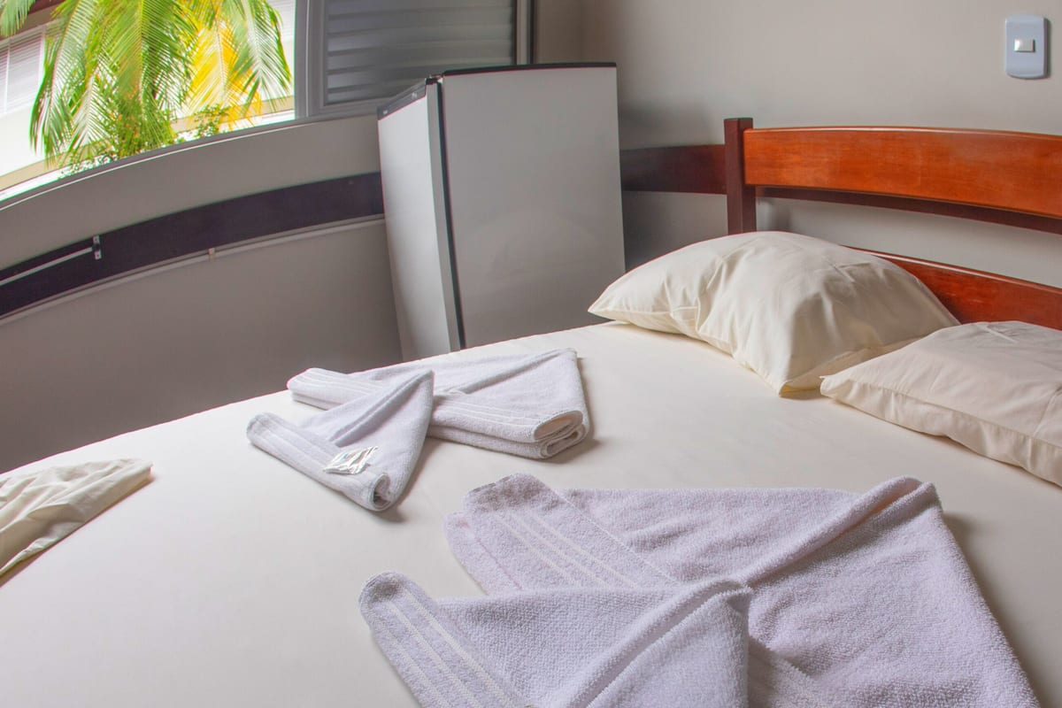 Cama bem arrumada com almofadas e toalhas dobradas, cabeceira de madeira, parede branca e janela com vista para palmeiras.