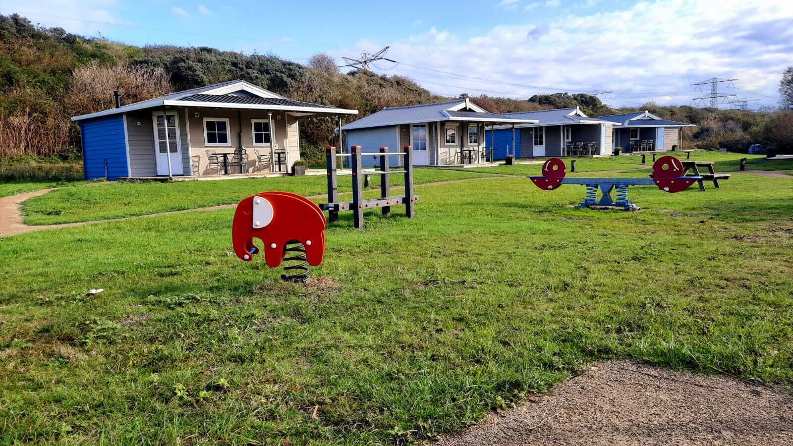Une aire de jeux avec une structure en forme d’éléphant rouge, un bâtiment bleu doté d’un patio couvert et une table de pique-nique dans un espace verdoyant.