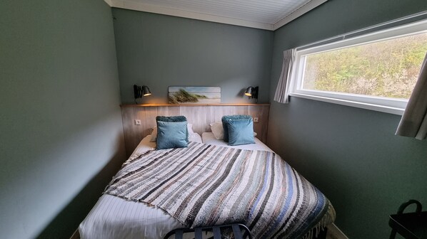 A bedroom with a bed, two pillows, a bedside table, a lamp, a window, and a view of greenery outside.