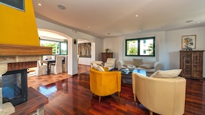 A spacious living area with a fireplace, yellow and white furniture, and large windows offering views of greenery.
