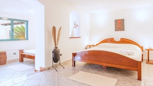A bedroom with a wooden bed, a bedside table, a nightstand, a vase with dried plants, and a window with a view of palm trees.