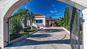 A paved driveway leading to a house with a red-tiled roof, surrounded by greenery and palm trees.