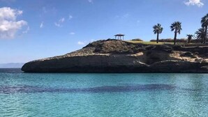 Una vista despejada de una playa con aguas turquesas, un afloramiento rocoso y una pequeña construcción en la cima.
