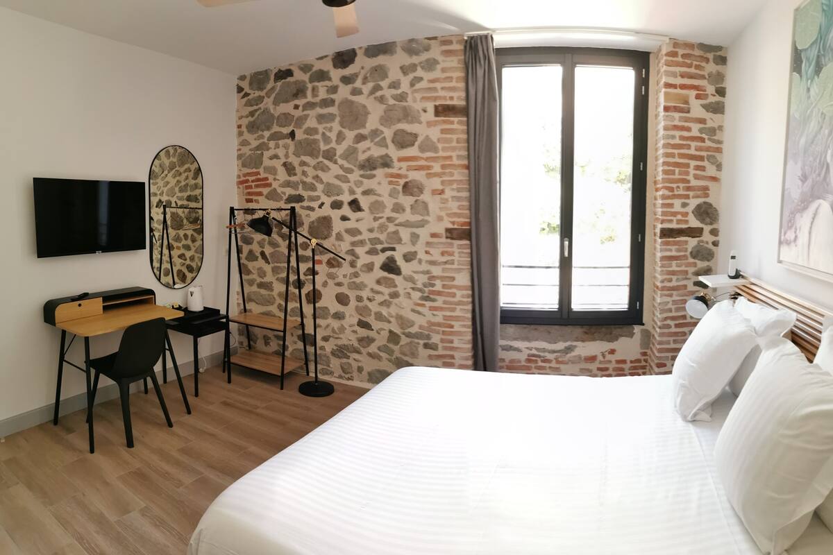 A bedroom with a stone wall, a bed with white bedding, a desk with a chair, and a television on the wall.