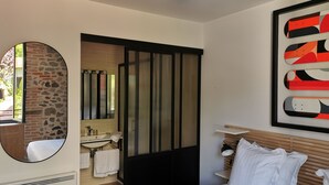 Una habitación de hotel moderna con una cama, un espejo redondo, un baño con lavabo y ducha, y un cuadro abstracto grande en la pared.