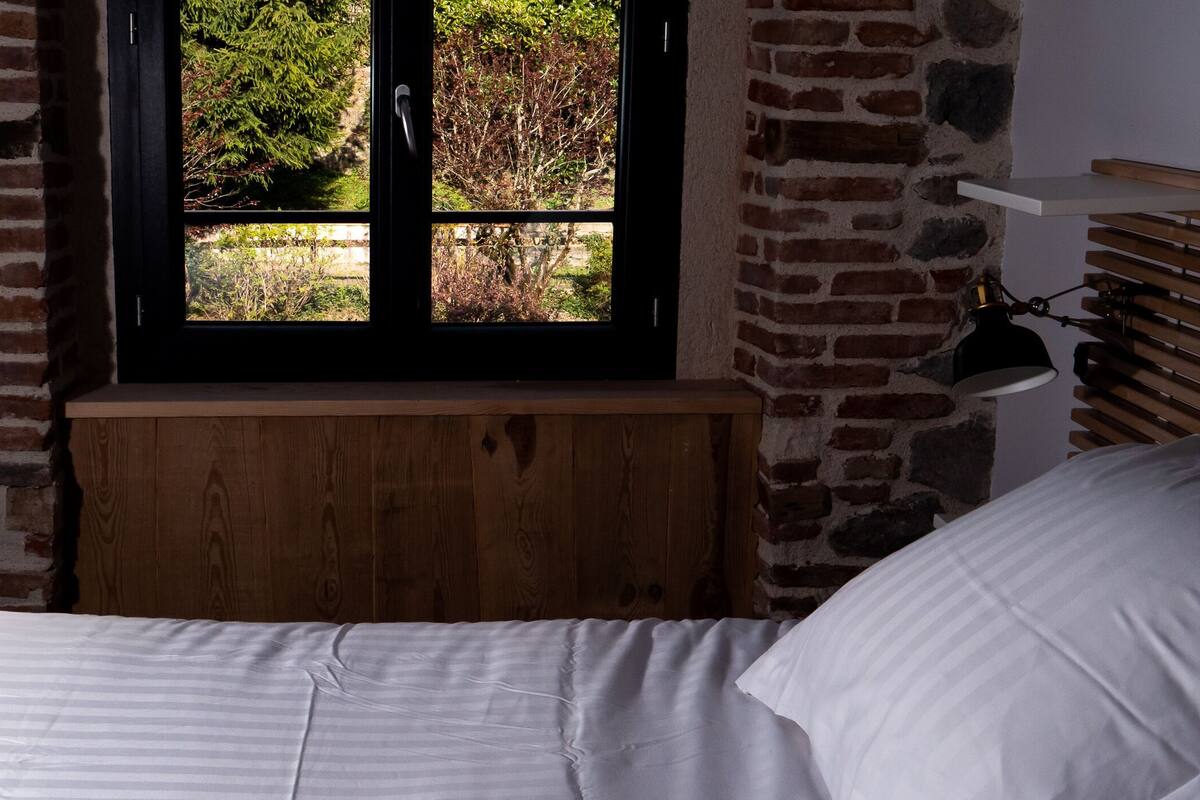 A room with a bed, a window with a view of greenery, a brick wall, and a lamp on a bedside table.