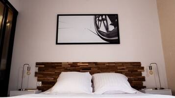 Une chambre d’hôtel avec une tête de lit en bois, un lit recouvert de draps blancs, deux tables de chevet avec des lampes et une œuvre d’art abstraite encadrée accrochée au mur.