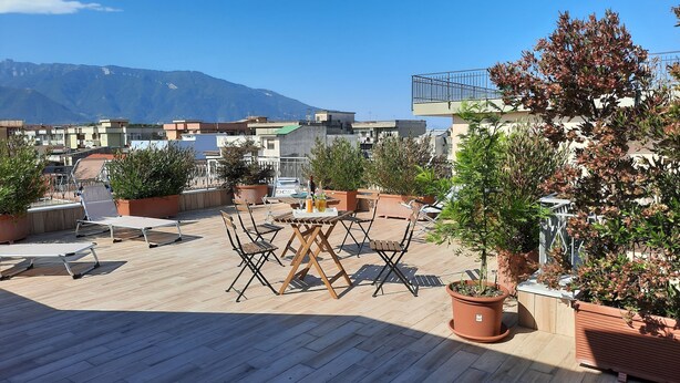 A rooftop terrace with wooden flooring, outdoor furniture, potted plants, and a view of a mountain range.