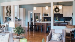 Cozinha moderna com armários brancos, eletrodomésticos de aço inoxidável e banquetas. Há uma área de jantar com mesa e cadeiras, e uma sala de estar com sofá e um banquinho verde.