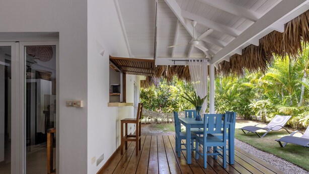 Un espace extérieur abrité, doté d’une terrasse en bois, d’une table avec des chaises et offrant une vue sur un jardin luxuriant agrémenté de palmiers.