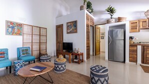 Un salon moderne comprenant un canapé, des poufs, une table basse et un téléviseur. Il y a également un coin cuisine avec un réfrigérateur et des placards en bois.