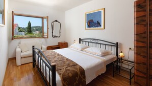 Een slaapkamer met een bed, een nachtkastje met een lamp, een raam met uitzicht op gebouwen en bomen, een stoel en een spiegel aan de muur.