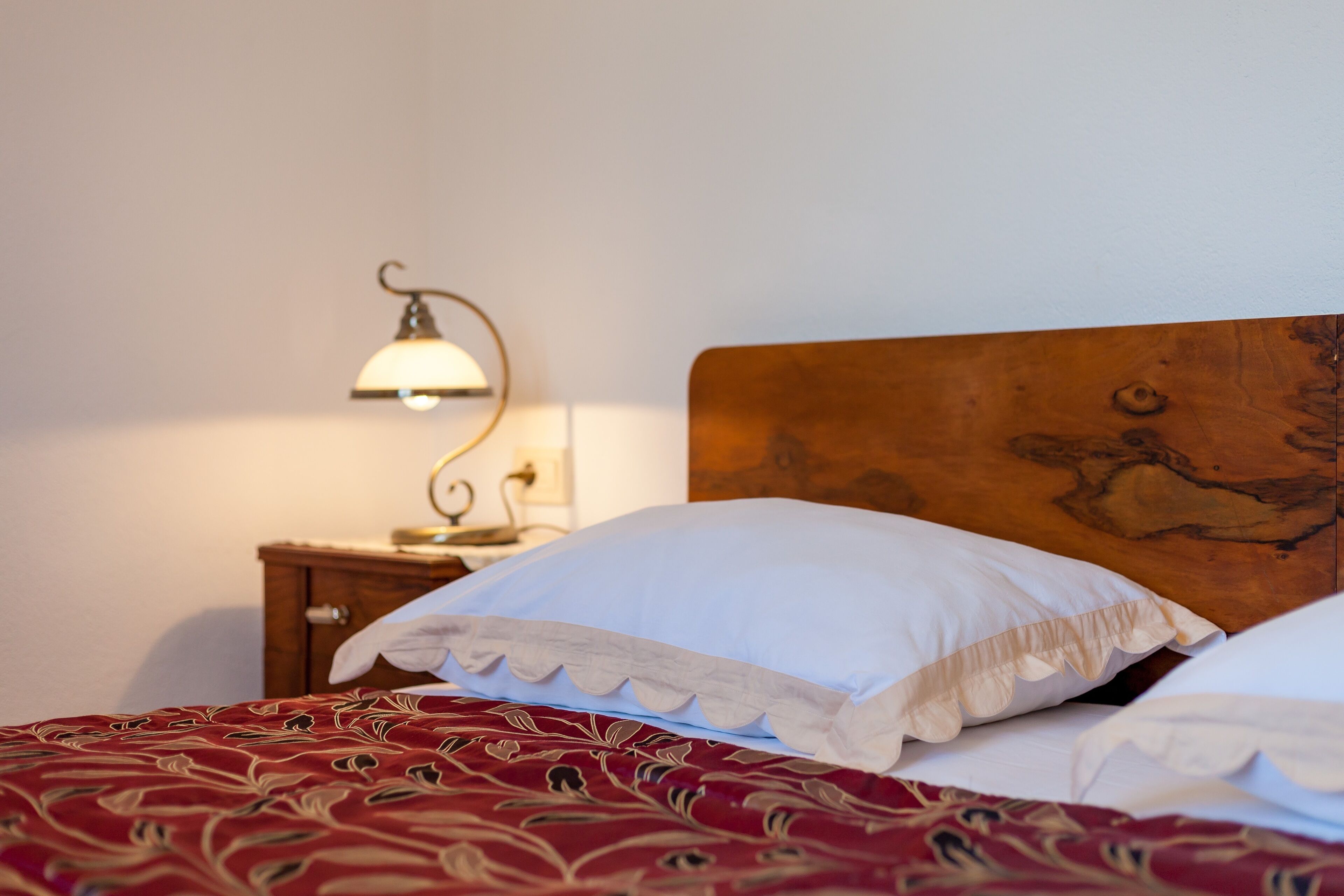 Un lit bien fait, avec une couvre-lit à motifs rouges et des oreillers blancs, une tête de lit en bois et une table de chevet surmontée d’une lampe.