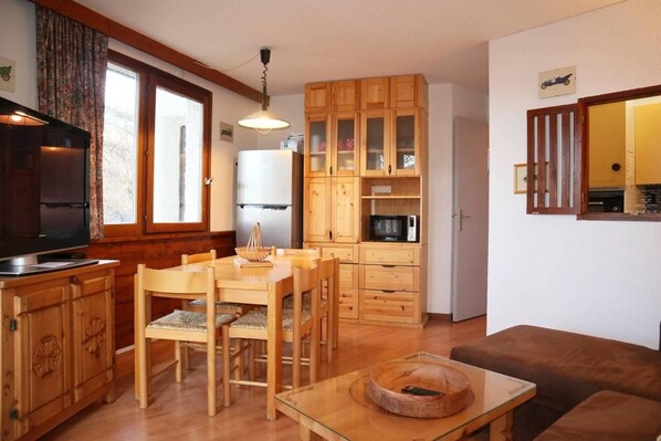 A compact kitchen with wooden cabinets, a dining table with chairs, a television, and a refrigerator.