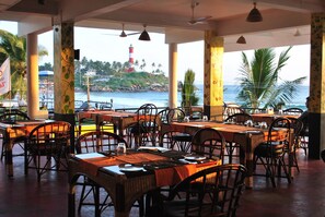 An outdoor restaurant with wicker tables and chairs, a view of the ocean, and a lighthouse in the background. - Newton Neelakanta Boutique – Kovalam (Neyyattinkara)