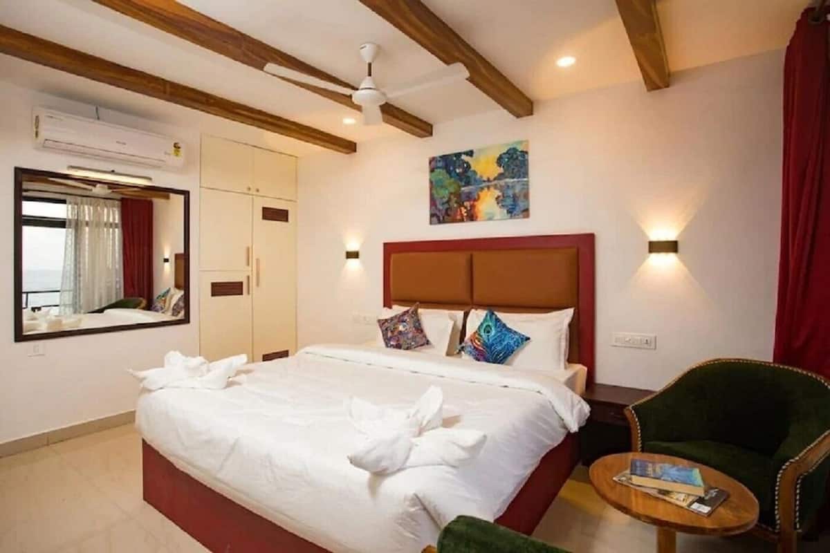 A hotel room with a large bed, a mirror, an air conditioning unit, a ceiling fan, a painting, and a green armchair.