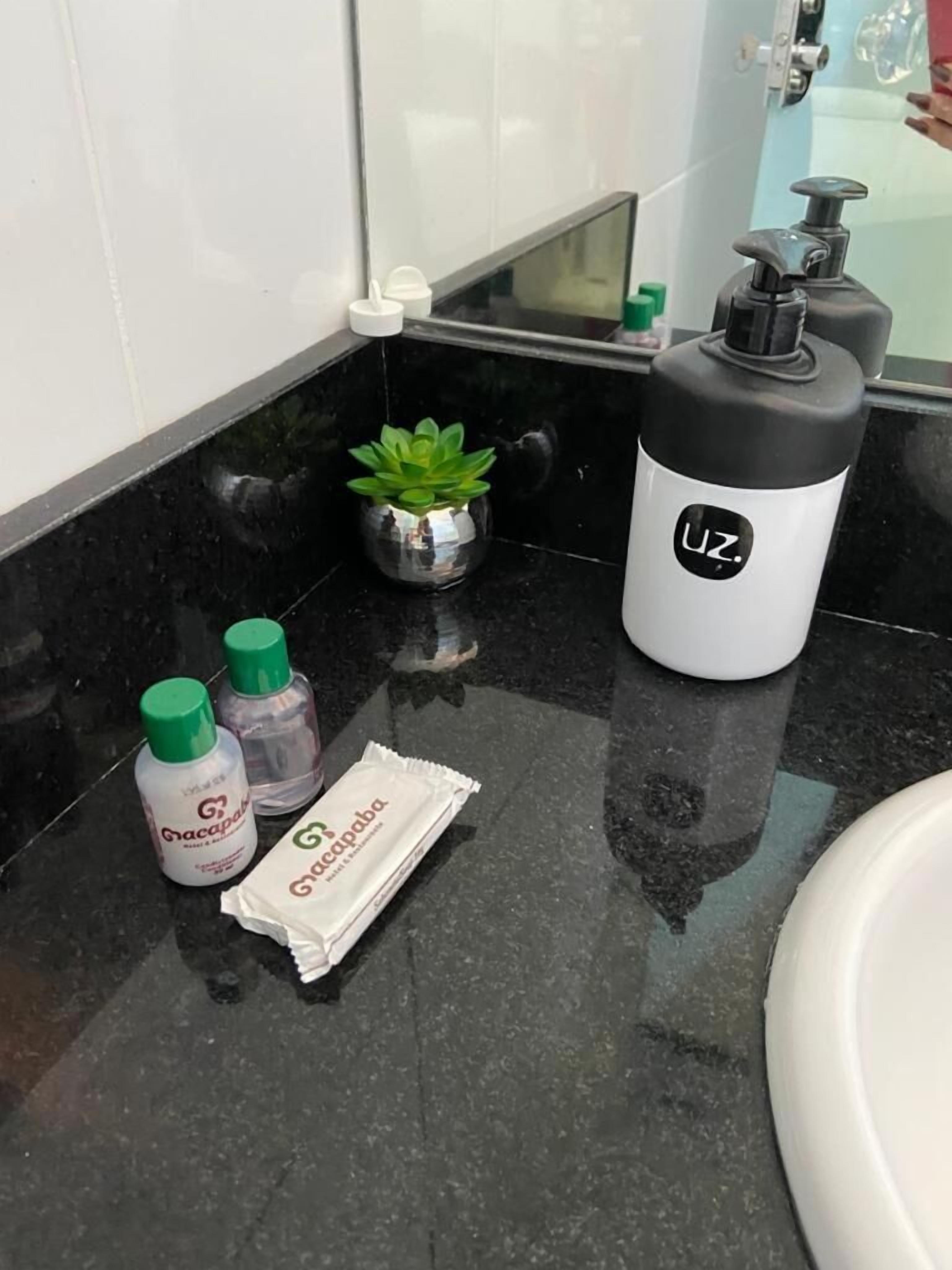 Un baño con encimera negra, una pequeña planta en maceta, una botella de Gracapab, un paquete de Gracapab y una botella de agua blanco y negro con el logo 'UZ'.