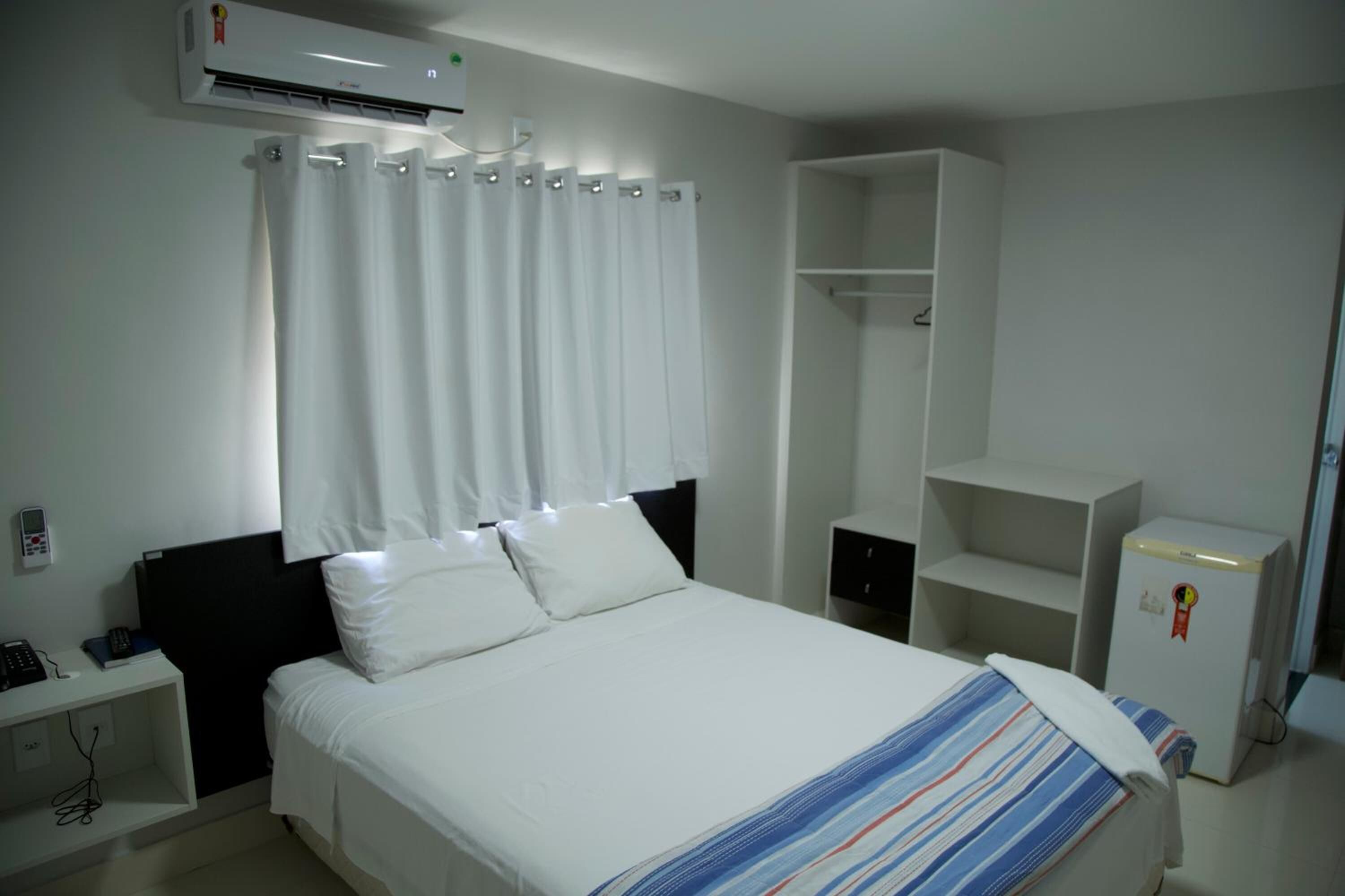 A hotel room with a bed, white curtains, a wardrobe, a desk with a phone, and a small refrigerator.