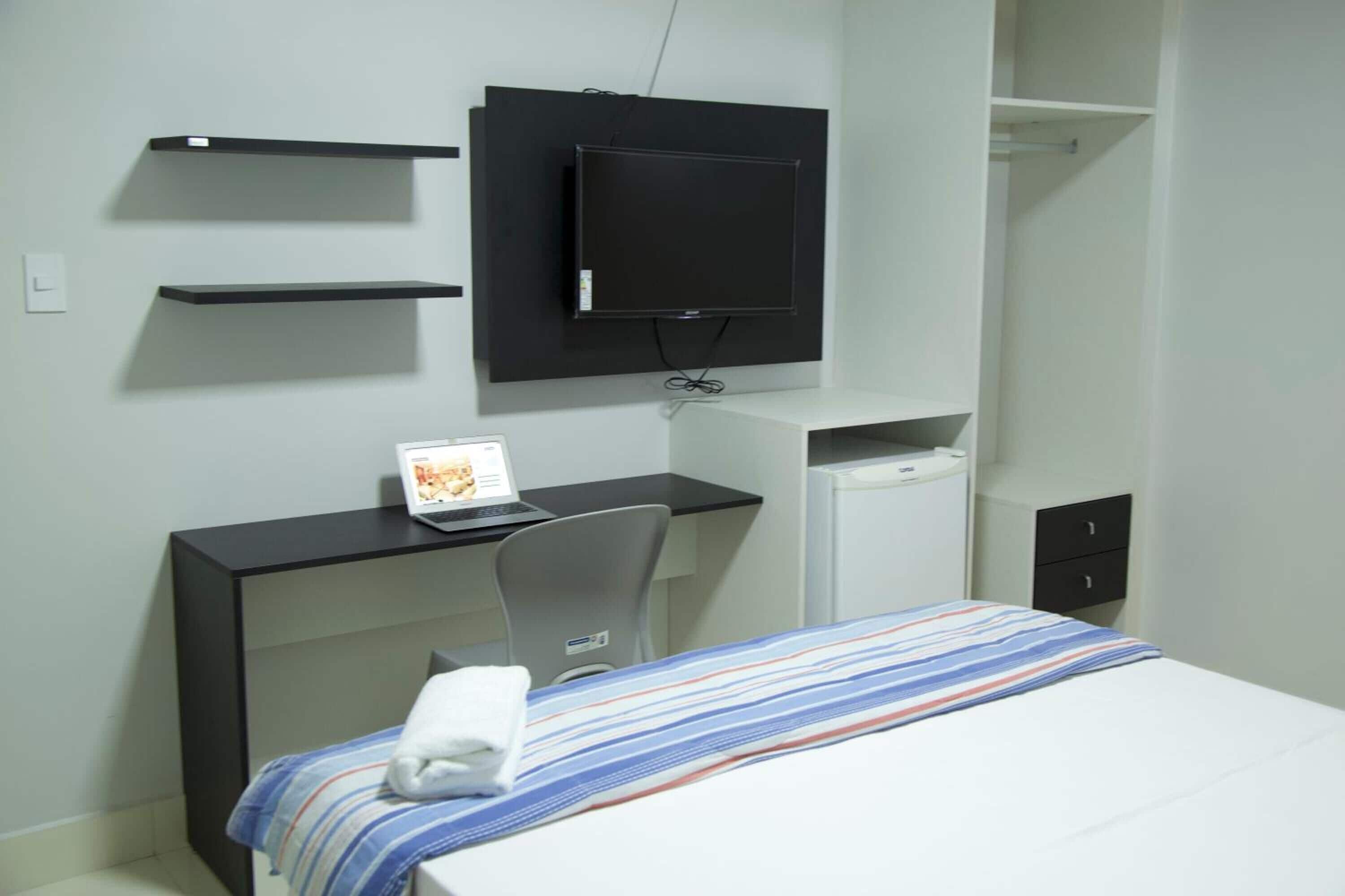 A hotel room with a bed, a desk with a laptop, a TV mounted on the wall, and a small cabinet.