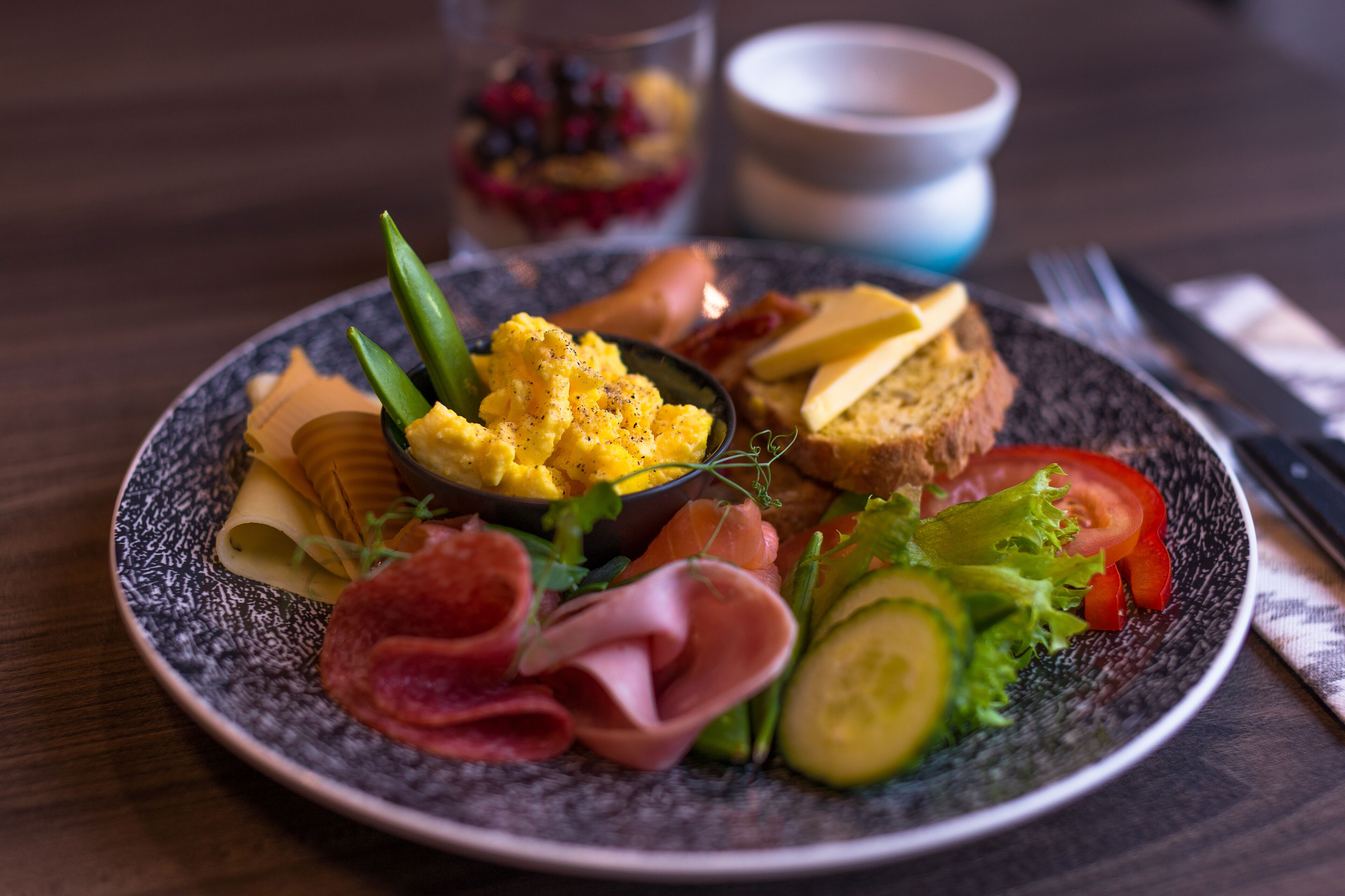 A plate of food with scrambled eggs, smoked salmon, ham, cucumber, and tomato slices.