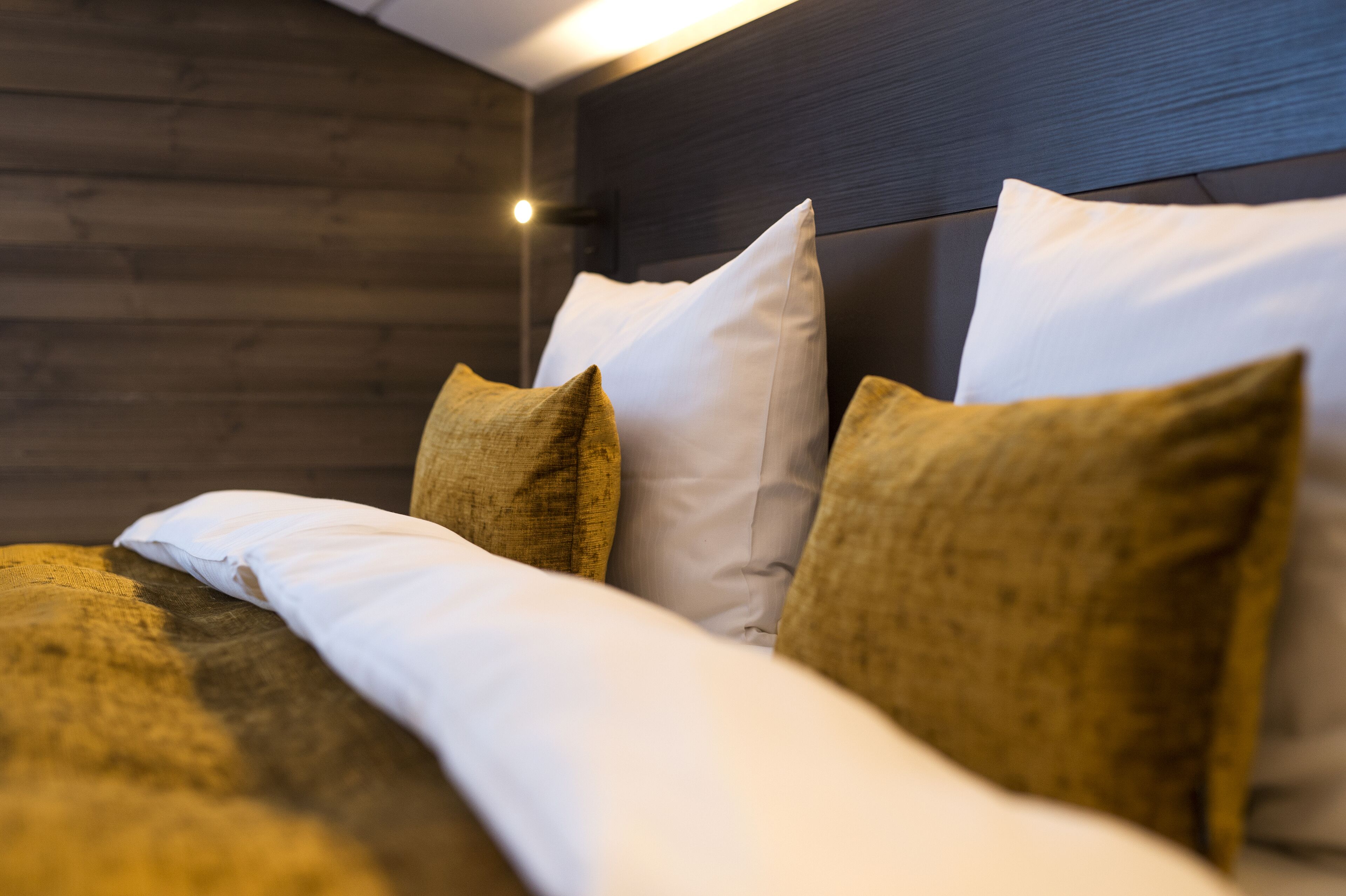 A neatly made bed with white and gold pillows, a wooden headboard, and a wall with a wooden panel design.