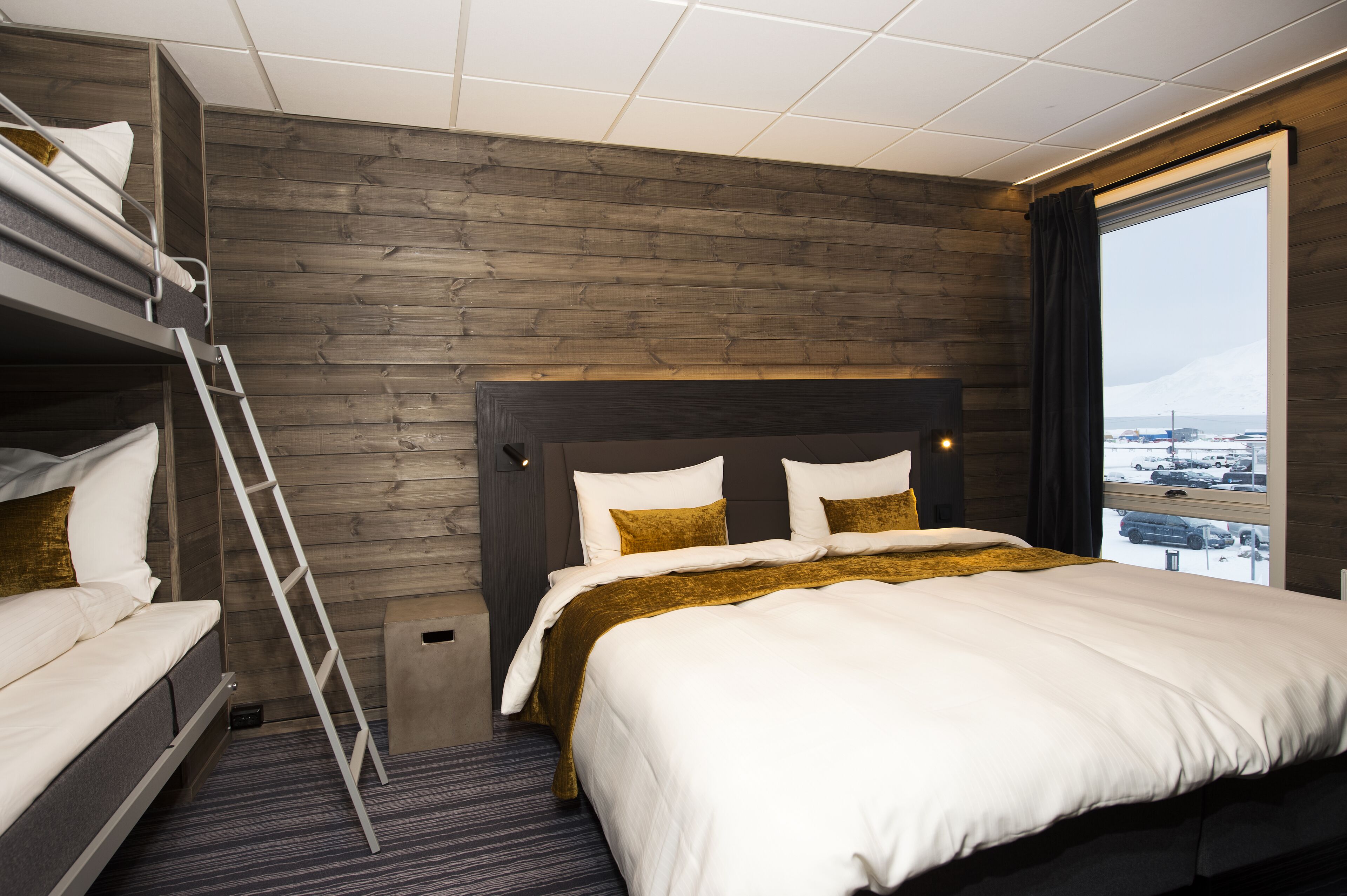 A hotel room with a bunk bed, a large bed, a wooden wall, a window with a view, and a ladder.