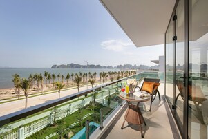 A balcony with a glass railing overlooking a beach and ocean. There is a table with chairs and a small garden area.