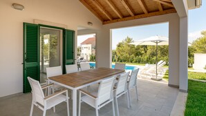 A covered patio with a wooden table and white chairs, overlooking a swimming pool and greenery. - Villa Makarac (Milna)