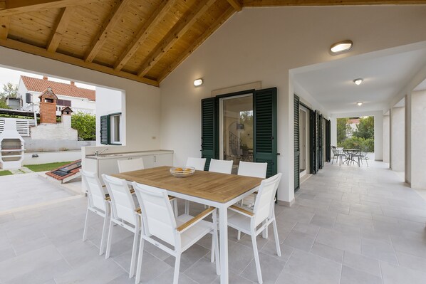 An outdoor patio with a wooden table and white chairs, a brick oven, and a view of a house in the background.