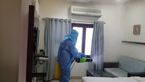 Uma pessoa com equipamento de proteção usando um pulverizador em quarto de hotel, com uma cama, uma mesinha e uma janela com cortinas.