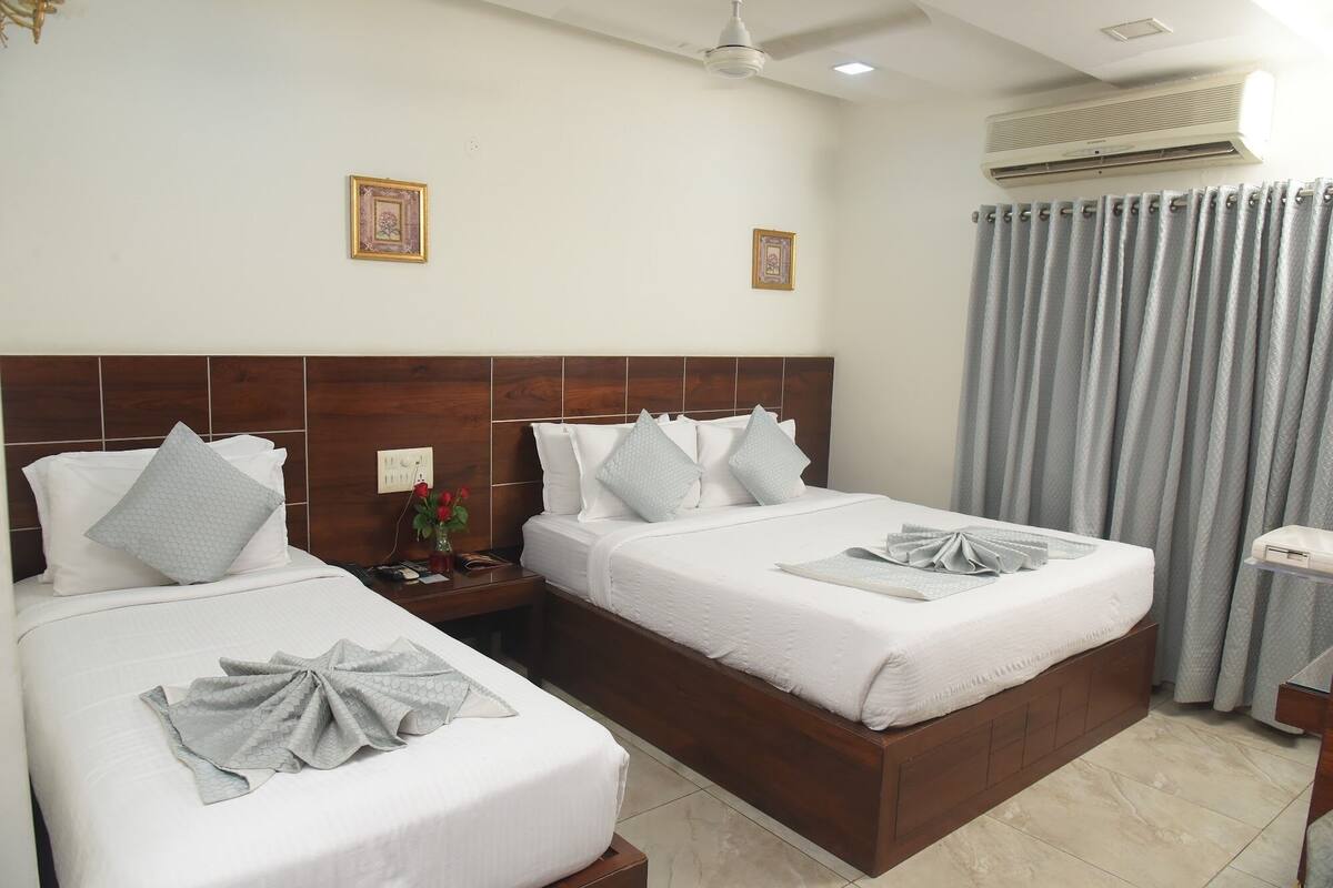 Habitación de hotel con dos camas, cabecera de madera, mesita de noche, un jarrón con flores y una ventana con cortinas.