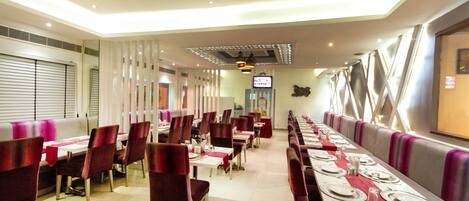 A restaurant with long tables, red chairs, and a TV screen on the wall.