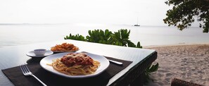 Uma mesa de jantar com pratos de espaguete e batatas fritas, montada numa praia com vista para o mar e um veleiro ao longe.